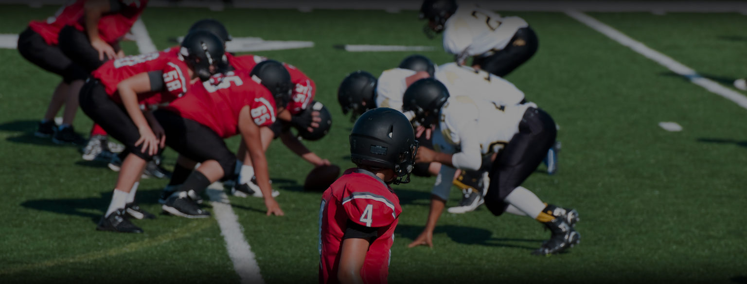 High-angle action view of youth football teams lined up at the line of scrimmage on a green turf field.