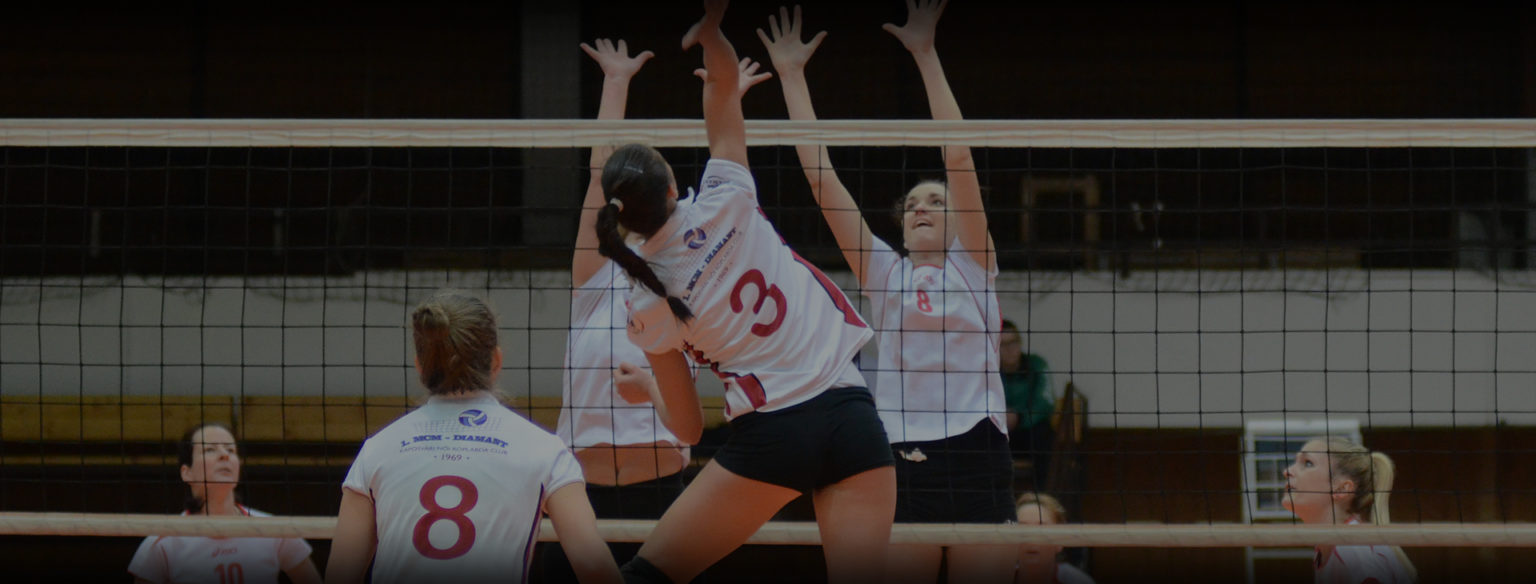 Women's volleyball team wearing custom white and red jerseys jumping to block a spike at the net.