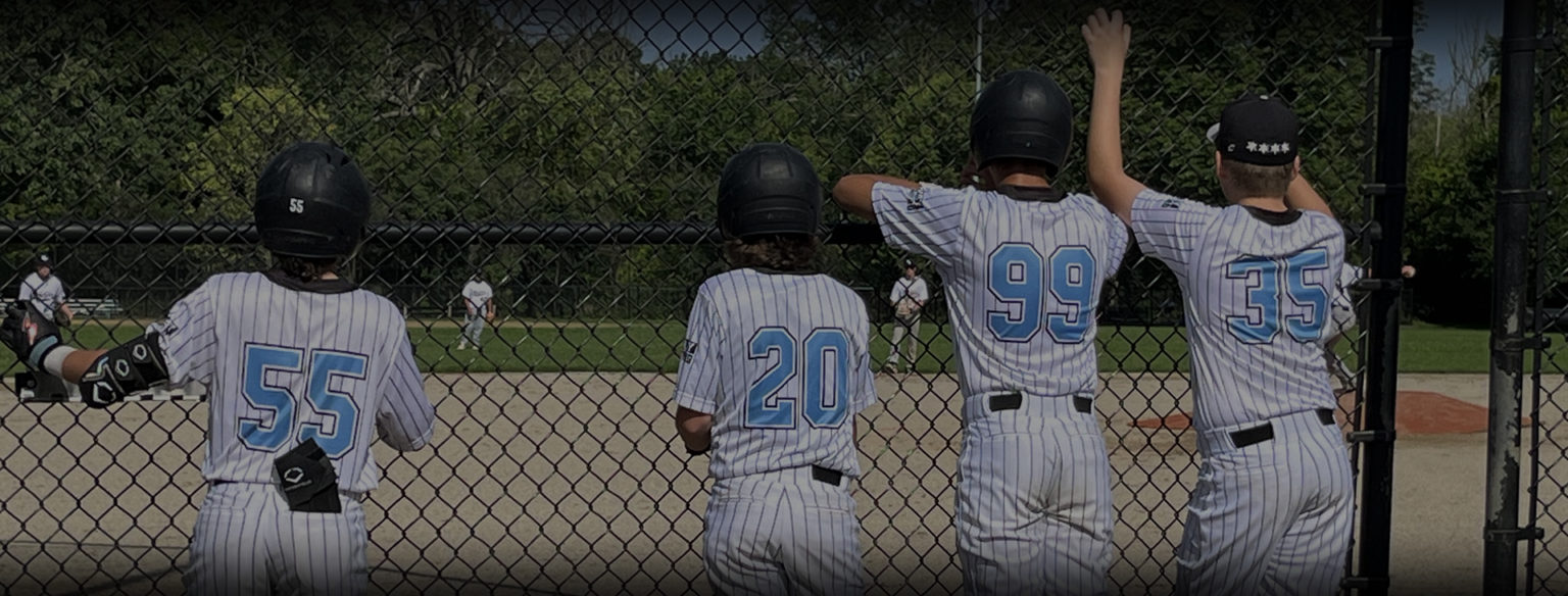 Back view of four youth baseball players in pinstripe custom jerseys watching the game through a chain-link fence dugout.